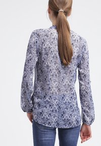 Blouse à manches longues en tissu léger et texturé avec un motif moucheté bleu et blanc. Présente un ourlet arrondi et des poignets boutonnés.