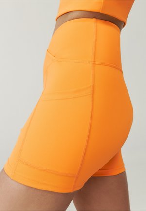 Close-up van een persoon die feloranje sportshorts met hoge taille en een zijzak draagt, met gedetailleerde stiksels en textuur van de stof zichtbaar.