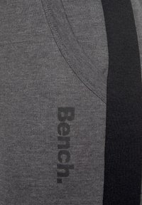 Graue Stoff-Jogginghosen mit schwarzem Streifen an der Seite, ausgestattet mit einer Tasche und dem Wort "Bench." in schwarzer Schrift, mit strukturiertem und glattem Finish.