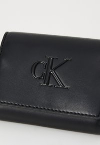 Juodas odinis piniginė su blizgiu iškilmingu "CK" logotipu, matomomis siūlėmis ir kompaktišku, stačiakampiu dizainu.