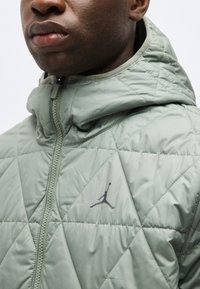 Κοντινό πλάνο ενός ατόμου που φοράει ανοιχτό γκρι μπουφάν με καπιτονέ επένδυση, κουκούλα και το λογότυπο Jumpman σε μαύρο χρώμα στο στήθος.