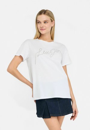 T-shirt blanc à manches courtes arborant un design en pierres précieuses argentées épelant "Lujo" sur le devant, associé à une jupe en jean.