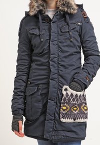 Marinblå parka med fuskpäls, fickor med dragkedja och knappar, ribbade ärmslut och dekorerad mönstrad ficka med gula och grå inslag.