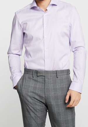 Man draagt een lichtpaarse knoopjes overhemd in zijn grijze geruite nette broek gestopt, met handen in de zakken, staand tegen een effen achtergrond.