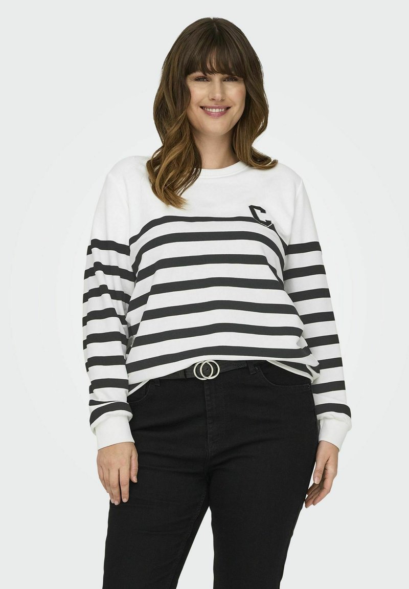 Sudadera blanca con rayas horizontales negras, cuello redondo y un logo en el pecho. Combinada con pantalones negros, que tienen una hebilla de cinturón circular.