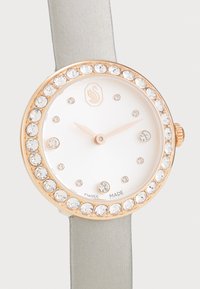 Orologio in oro rosa con quadrante bianco, dettagli in cristallo e cinturino in satin grigio chiaro. Presenta un design semplice con indicatori delle ore minimalisti.