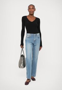 Frau mit rasiertem Kopf, die ein schwarzes Langarmoberteil, hellblaue Jeans und burgunderfarbene Ballerinas trägt und eine metallisch geflochtene Handtasche hält.