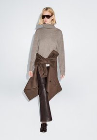 Maglione grigio a collo alto abbinato a un avvolgente mantello di lana marrone, pantaloni attillati marroni scuri e mocassini neri. Il modello indossa occhiali da sole oversized.