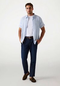 Lyseblå kortærmet skjorte med knapper over en hvid T-shirt, kombineret med mørkeblå bukser og brune slip-on sko. Casual pasform og rent design.