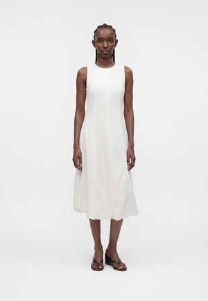 Femme debout, portant une robe midi blanche sans manches et des sandales compensées noires, sur un fond uni gris clair.