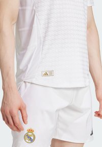 Camiseta atlética blanca con patrón texturizado, con la marca y logo de Adidas. Combinada con pantalones cortos blancos que exhiben el emblema del Real Madrid.