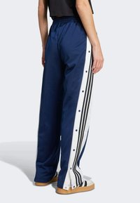 Person wearing navy blue track pants with white side stripes, black vertical stripes, and snap buttons, paired with white sneakers with black stripes.