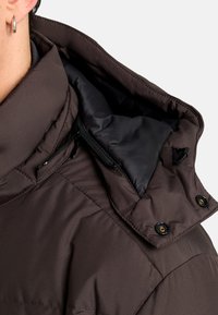 Giacca a vento marrone con colletto alto, dotata di una fodera nera, tasca con cerniera e bottoni a pressione. Texture liscia e design trapuntato.