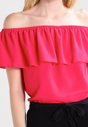 Kvinde iført en lyserød off-shoulder top med flæsekant og sorte bukser bundet med sløjfe i taljen.
