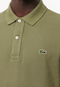 Olivgrön polo t-shirt med en strukturerad tyg, två knappar vid kragen och en liten broderad krokodil-logga på vänster bröst.