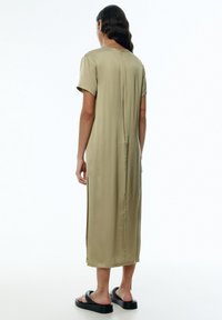 Knielanges, unifarbenes beige Kleid aus glattem Stoff, mit kurzen Ärmeln, Seitenschlitzen und einem Knopfverschluss auf der Rückseite. Kombiniert mit schwarzen Sandalen.