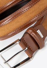 Bruine suède en leren riem met contrasterende stiksels. Voorzien van een zilverkleurige metalen gesp en een gegraveerde logoplaat op de band.