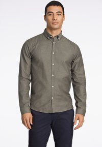 Camisa abotonada en verde oliva, con textura tejida, mangas largas, cuello estructurado y botones blancos. Combinada con pantalones azul marino.