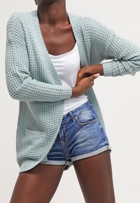 Cárdigan de punto azul claro con delantero abierto, mangas largas y dos bolsillos frontales, combinado con pantalones cortos de mezclilla casuales.