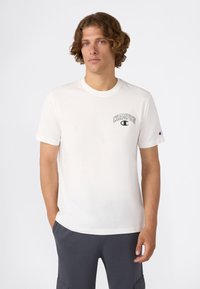 Hombre joven con cabello rizado, vistiendo una camiseta blanca de Champion y pantalones gris oscuro, de pie con una mano en el bolsillo frente a un fondo liso.