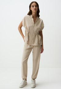 Maglione beige con zip, maniche corte, vita cintrata e due tasche frontali, abbinato a pantaloni con polsini elasticizzati e scarpe bianche.