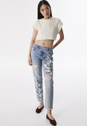 Witte gebreide crop top, lichtblauwe distressed jeans met zwarte tekst en scheuren, en bruine flats met kettingaccenten. Eenvoudig en casual ontwerp.
