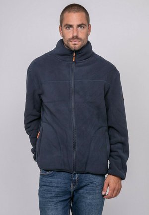 Homme de face portant une veste polaire bleu foncé zippée à col montant et un jean bleu, debout devant un fond clair et uni.