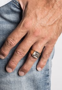 Kvadratinės formos žiedas su juodu ir baltu akmeniu auksinės spalvos metaliniame laikiklyje, dėvimas ant rankos prieš džinsinio audinio foną.