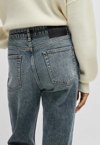 Jeans de mezclilla azul claro con cintura alta, bolsillos traseros prominentes y cinturilla de marca negra. Tejido texturizado con costuras visibles.