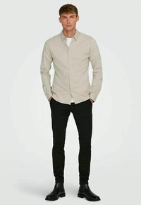 Homem em pé com as mãos nos bolsos, vestindo uma camisa bege de botão, uma t-shirt branca por baixo, calças pretas e botas de cano baixo pretas, em fundo branco.