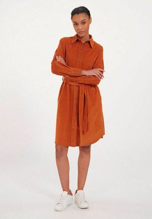 Robe en velours côtelé couleur rouille avec fermeture à boutons, ceinture à la taille, manches longues et col. Portée avec des baskets blanches. Texture lisse.