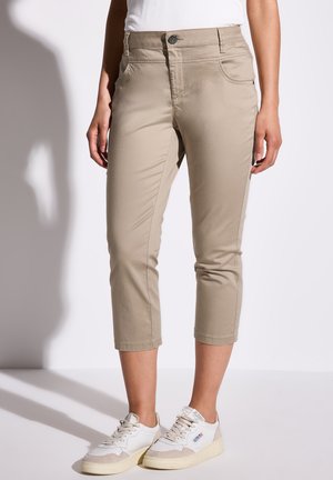 Beige cropped bukser med lynlås og knaplukning, der har forlommer og en tætsiddende pasform. Sammen med hvide og beige sneakers.