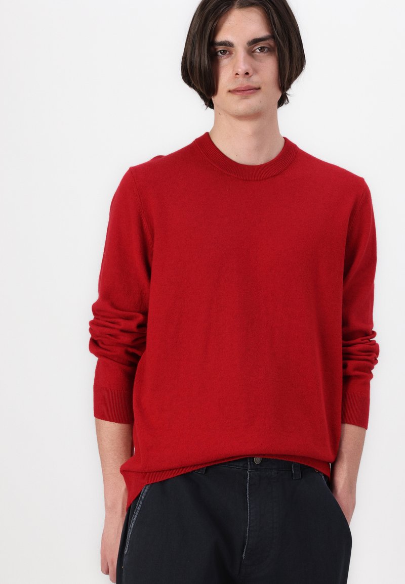 Suéter de punto rojo con cuello redondo, mangas largas y puños acanalados. La tela parece suave y lisa con un corte relajado. Combinado con pantalones oscuros.