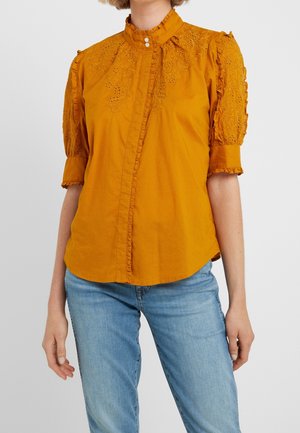 Kvinde iført sennepsgul bluse med broderet blomsterblonde og flæsekant, kombineret med lyseblå jeans, stående foran en hvid baggrund.