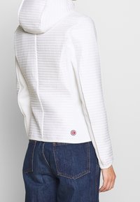 Vit quiltad jacka med huva, med en texturerad yta och en liten rund logotyp på nedre ryggen. Parat med mörkblå jeans.