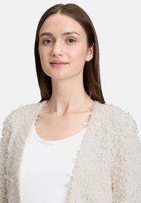 Cardigan en laine bouclée avec un design à ouverture frontale, tissu texturé et bords bruts, porté par-dessus une chemise blanche unie.