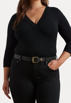 Kobieta ubrana w dopasowaną czarną bluzkę wiązaną z rękawami o długości ¾, czarne spodnie oraz czarny skórzany pasek z pozłacaną klamrą i logo RLL.