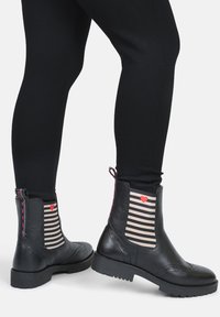 Stivaletti neri in pelle con lati elastici a strisce, dettaglio a forma di cuore rosa e superficie texturizzata, abbinati a leggings neri.