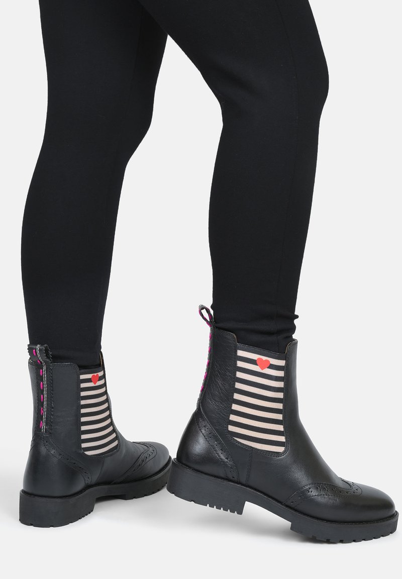 Stivaletti neri in pelle con lati elastici a strisce, dettaglio a forma di cuore rosa e superficie texturizzata, abbinati a leggings neri.