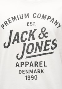 Κείμενο σε λευκό ύφασμα γράφει "Premium Company Est. Jack & Jones Apparel Denmark 1990" με φθαρμένη μαύρη γραμματοσειρά.