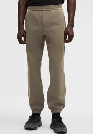 Beżowe spodnie dresowe z elastycznym pasem, bocznymi kieszeniami i zwężanymi ściągaczami, wykonane z gładkiego, elastycznego materiału. Zestawione z czarnymi trampkami.