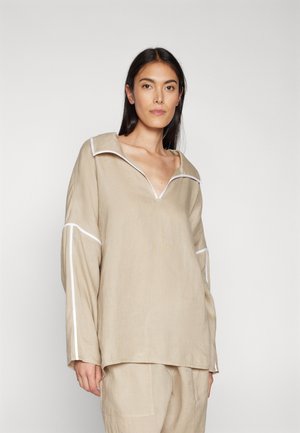 Haut en lin beige avec un large col, des détails en piping blanc et des manches longues, présente une coupe décontractée et une surface texturée.