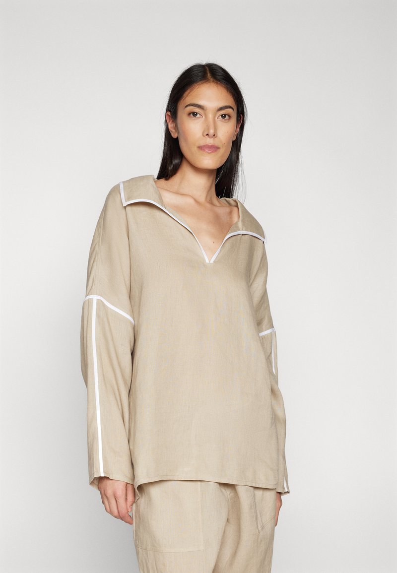Haut en lin beige avec un large col, des détails en piping blanc et des manches longues, présente une coupe décontractée et une surface texturée.