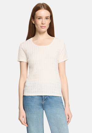 Jeune femme aux cheveux roux lisses portant un haut blanc texturé à manches courtes et un jean bleu clair, sur un fond uni.