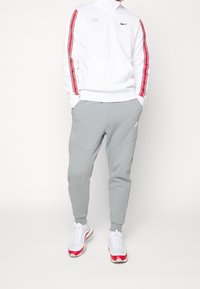 Vit zip-up sweatshirt med röda och svarta sidostripes, grå tapered joggers och vita sneakers med röda accenter.