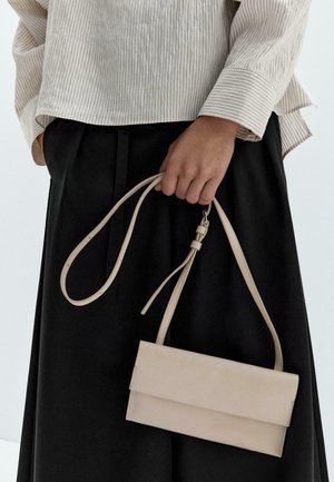 Beige leather crossbody bag with a smooth texture, fold-over design, and adjustable strap, held by a person in a striped top and black skirt.