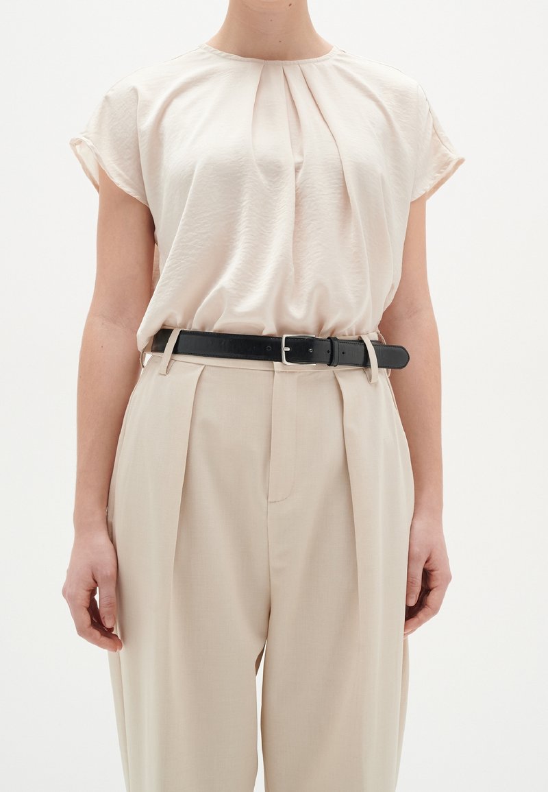 Lys beige bluse i blødt, tekstureret stof, med plisseringer ved halsudskæringen og korte ærmer, parret med lys beige plisserede bukser og et sort bælte.