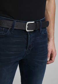 Cinturón de piel negra con una hebilla de metal plateado, que presenta un logo en relieve. Llevado con unos jeans de mezclilla oscuros, mostrando un primer plano del área de la cintura.