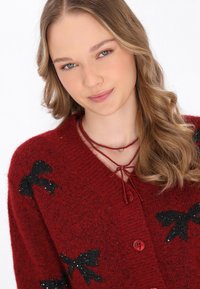 Röd fluffig cardigan med svarta paljettrosetter, med en v-ringning och röda knappar. Halsband med röda pärlor och en knytning.