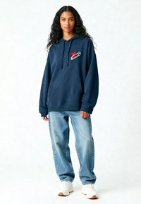 Felpa blu navy con tasca frontale, presenta una grafica con un cuore rosso e un fermaglio di sicurezza. Abbinata a jeans baggy azzurri e sneakers bianche.